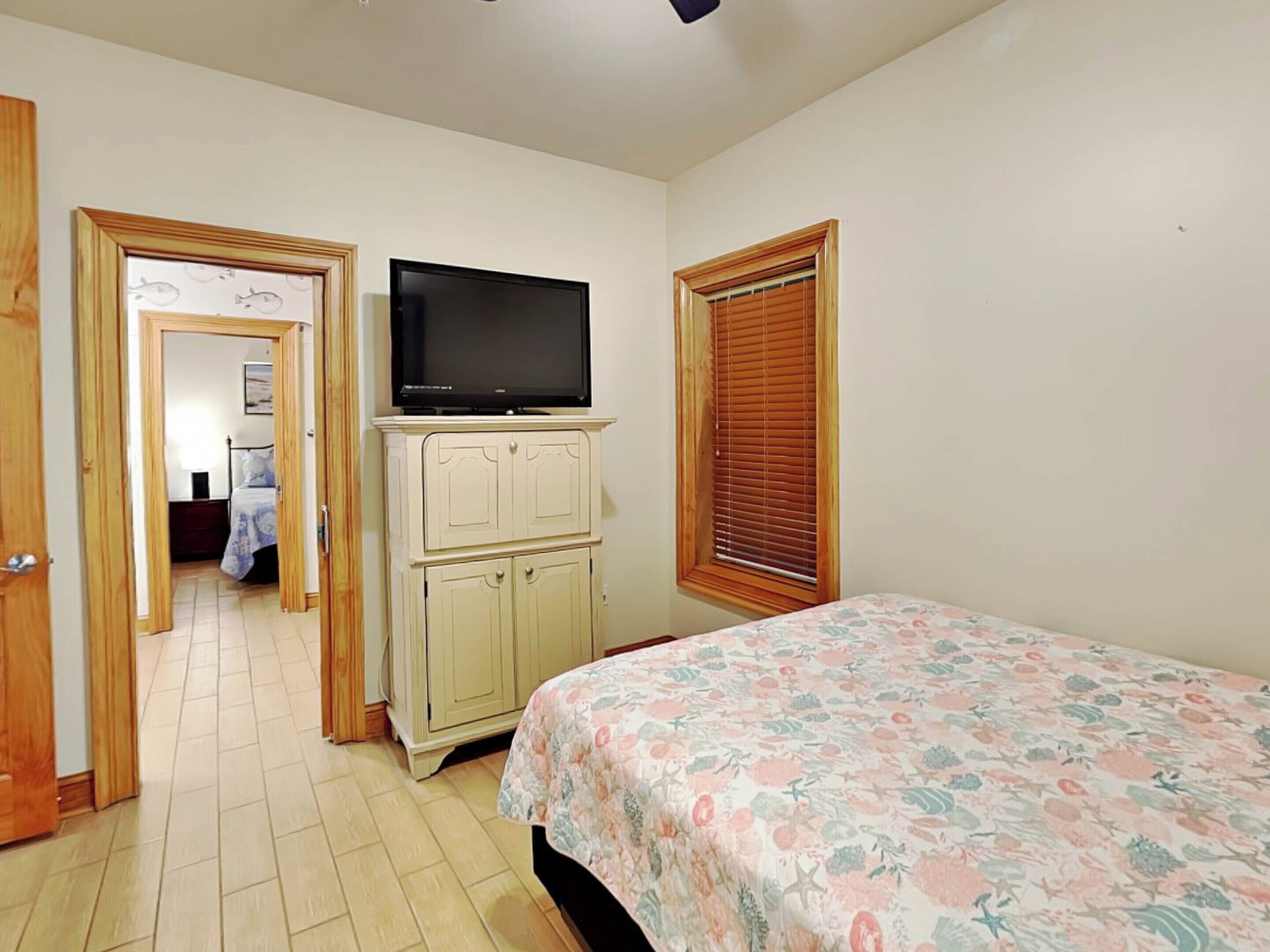 A bedroom with a bed, a television on a cabinet, a window with blinds, and a door leading to another room.