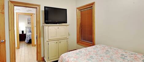 A bedroom with a bed, a television on a cabinet, a window with blinds, and a door leading to another room.