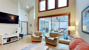 A spacious living room with a large flat-screen TV, leather sofas, a coffee table, and a large window overlooking a balcony. - The Gardens 703 by Tybee Vacation Rentals (Tybee Island)