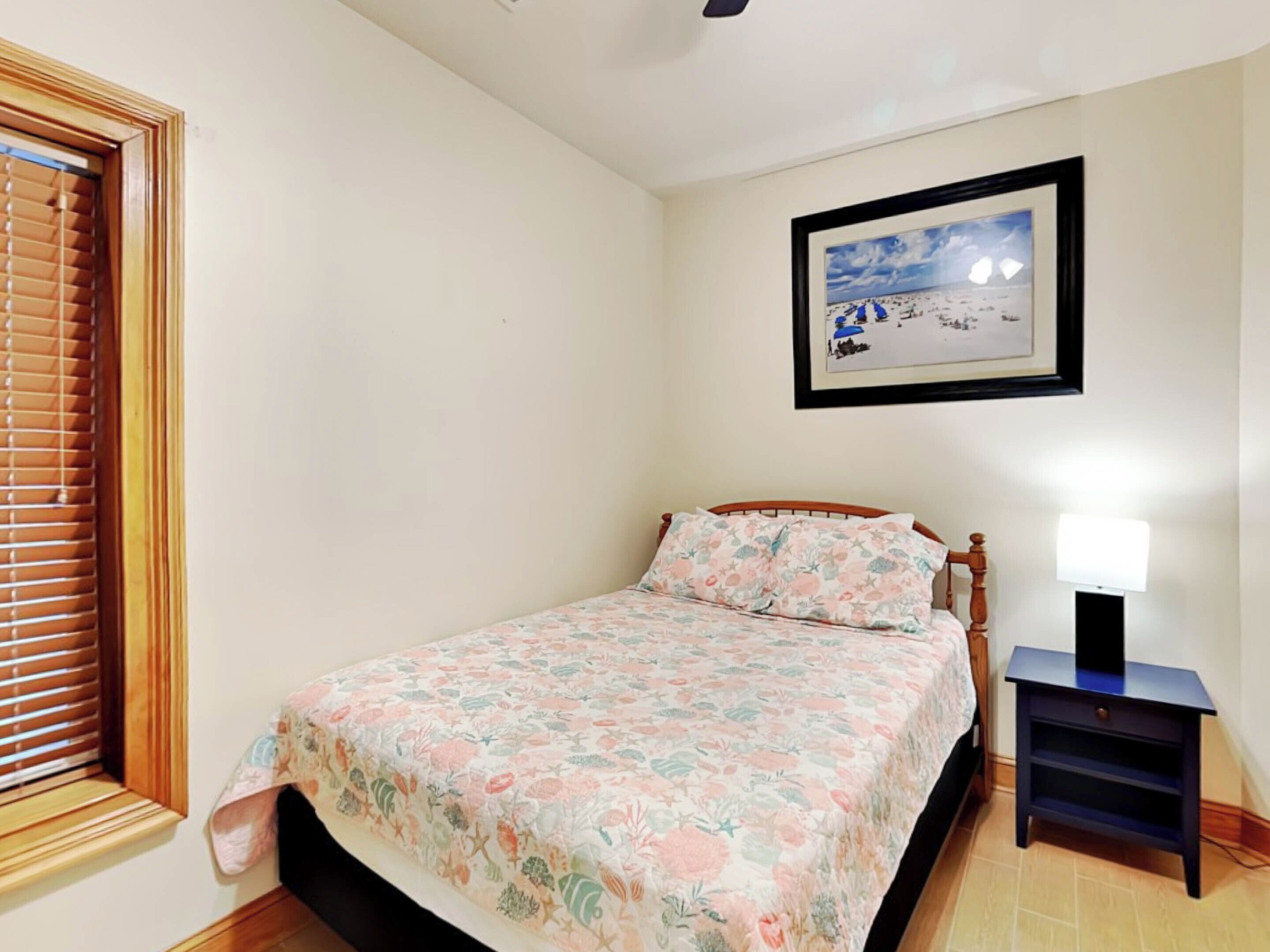 A bedroom with a bed, a window with blinds, a framed picture on the wall, and a bedside table with a lamp.