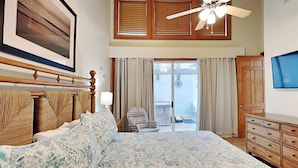 A bedroom with a large bed, a ceiling fan, a TV, and a balcony with a chair. - The Gardens 703 by Tybee Vacation Rentals (Tybee Island)