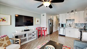 A modern living room with a flat-screen TV, a ceiling fan, and a dining area with red chairs. - Beach Racquet A209 by Tybee Vacation Rentals (Tybee Island)