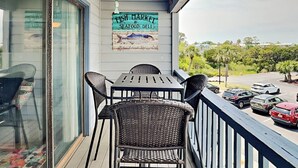 A balcony with a table and chairs, a sign for a fish market and seafood deli, and a view of parked cars and greenery. - Beach Racquet A209 by Tybee Vacation Rentals (Tybee Island)
