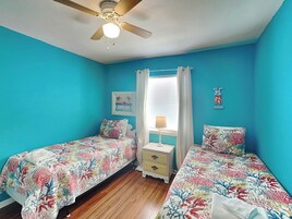 A room with two single beds, a ceiling fan, a window with curtains, a bedside table, and a wall with a 'VACATION' sign.