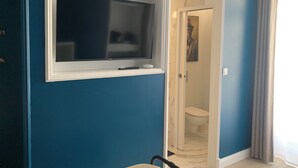 Een kamer met een aan een blauwe muur bevestigde televisie, een deur naar een badkamer en een raam met gordijnen.