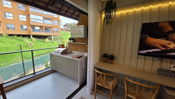 A balcony with a kitchen area, a TV, and a view of a building and greenery. - Flat Luxo e Conforto 224 Eco Resort by La Playa (Tamandaré)