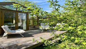 A wooden deck with two white lounge chairs and a glass-enclosed room with a sliding door. - Group Accommodation for 36 Guests (Scherpenzeel)