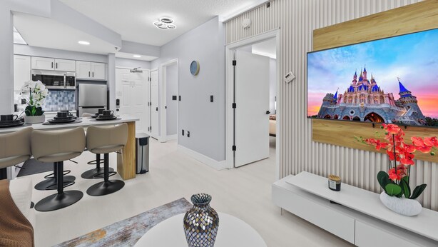 A modern kitchen with white cabinets, a microwave, and a refrigerator. There is a dining area with a white table and chairs. A large TV is mounted on the wall, displaying a castle.