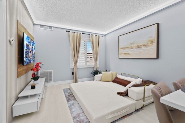 A modern bedroom with a large bed, a flat-screen TV, a bedside table with a vase of flowers, and a window with curtains.