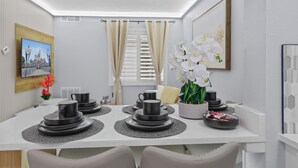 A dining area with a white table set for four, featuring black plates and cups, a vase with white flowers, and a framed picture on the wall.