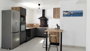 A modern kitchen with a built-in refrigerator, oven, and microwave. There is a wooden island with bar stools and a coffee machine. A large window provides a view of boats and a lighthouse. - New house, spacious, bright, quiet neighborhood in agglo, fenced. Garage (Plouharnel)