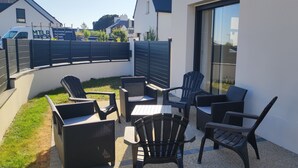 An outdoor patio area with wicker furniture and a table, surrounded by a white wall and a black fence. - New house, spacious, bright, quiet neighborhood in agglo, fenced. Garage (Plouharnel)