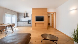 A modern living room with a flat-screen TV mounted on a wooden wall, a dining area with a table and chairs, and a kitchen in the background.