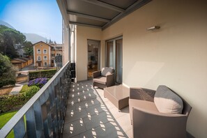 En balkon med sofa og lænestol, en glasdør der fører til et indre rum, og udsigt til omkringliggende bygninger.