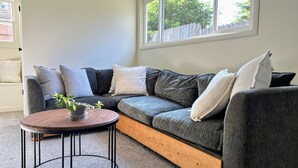 Een moderne woonkamer met een grijze modulaire bank, een houten salontafel en uitzicht op een witte schutting via een groot raam.