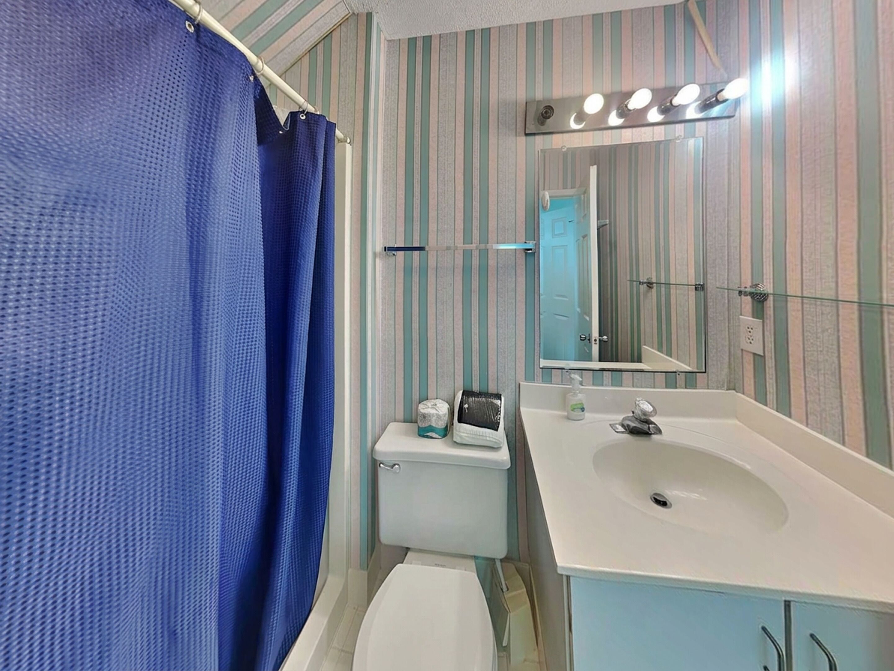 A bathroom with a striped wall, a white sink, a mirror with lights, a blue shower curtain, and a toilet.