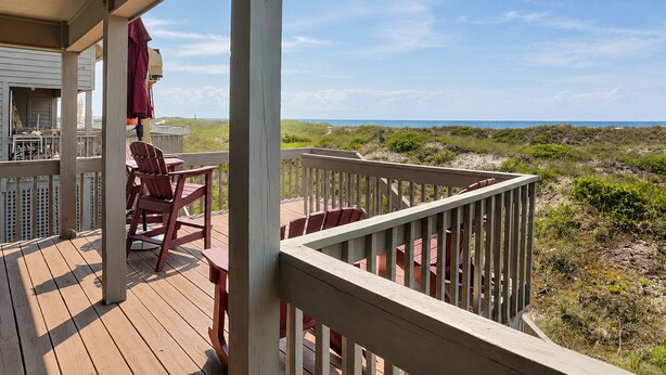 Een houten terras met stoelen en een tafel, uitkijkend op een strand met duinen en de oceaan.