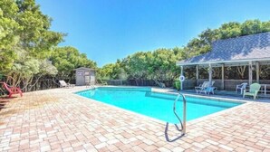 Een rechthoekig zwembad met een terras van baksteen, omgeven door bomen en een overdekt gedeelte met stoelen.