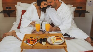 A couple in white robes sitting on a bed with a tray of breakfast items including toast, fruit, and juice. - Refúgio Serra e Flor (Urubici)