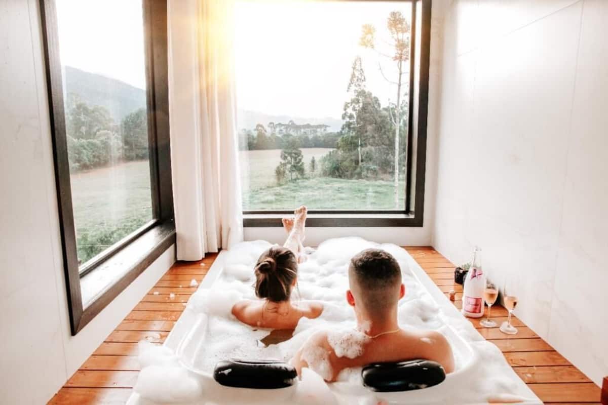 A room with a large window, a wooden floor, and a bathtub filled with bubbles.