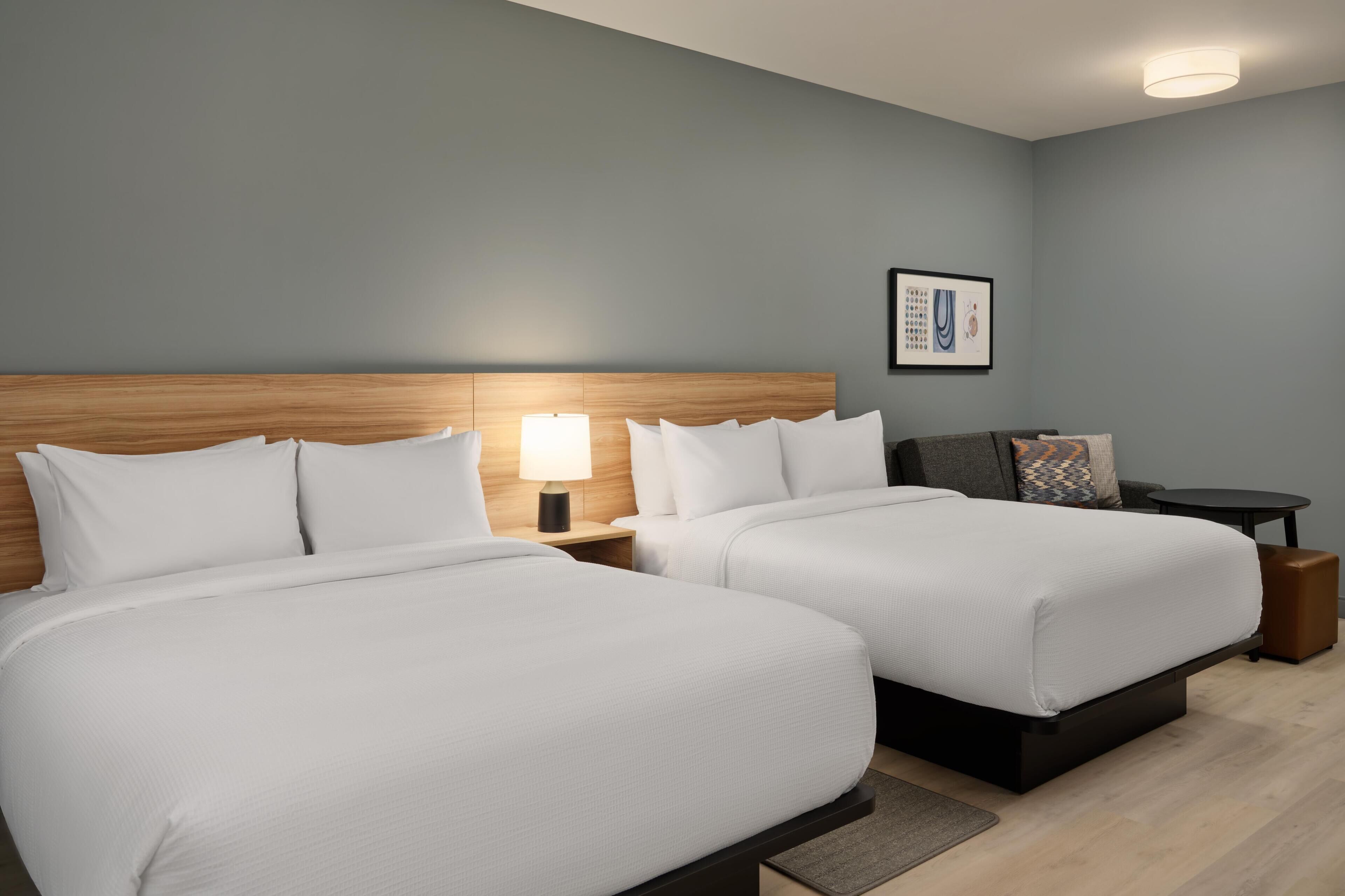 A hotel room with two beds, a nightstand with a lamp, a small table, and a framed picture on the wall.