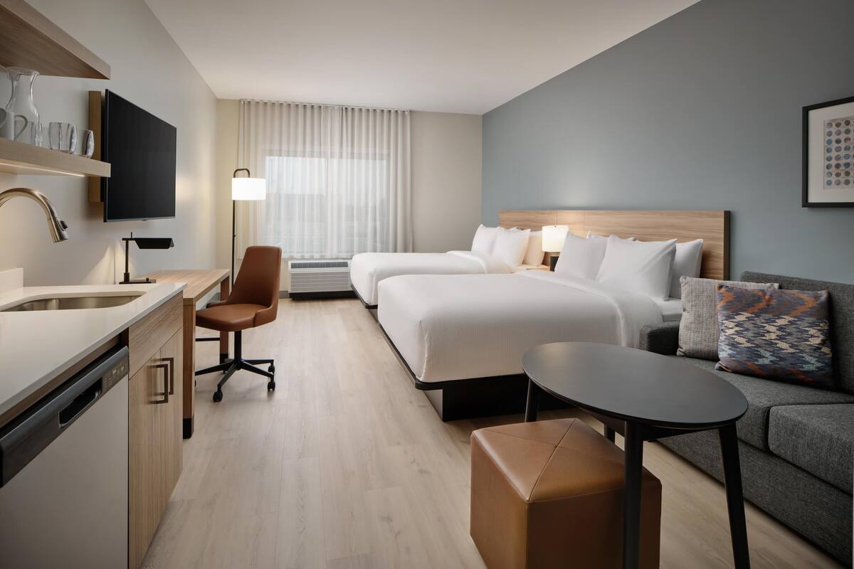 Une chambre d’hôtel avec un lit, un canapé, un bureau avec une lampe, une télévision et une fenêtre avec des rideaux.
