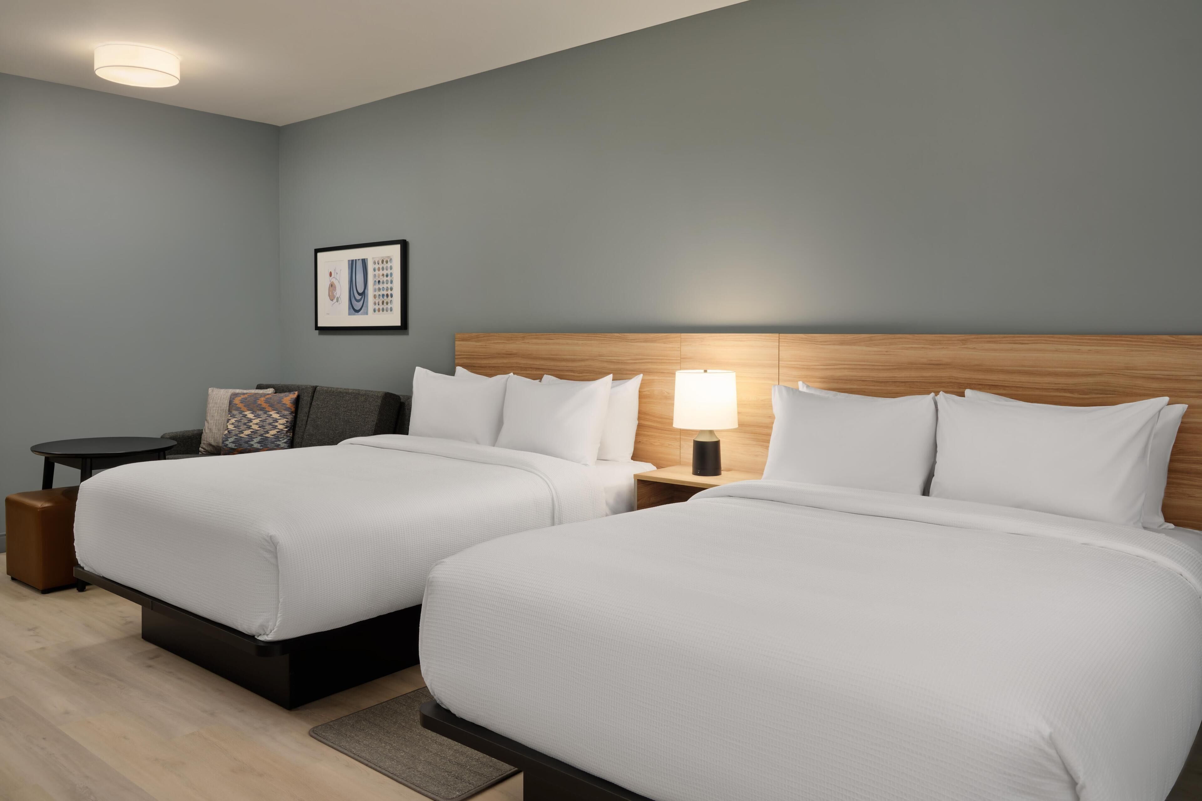 A hotel room with two beds, a small table, a nightstand, a lamp, and a framed picture on the wall.