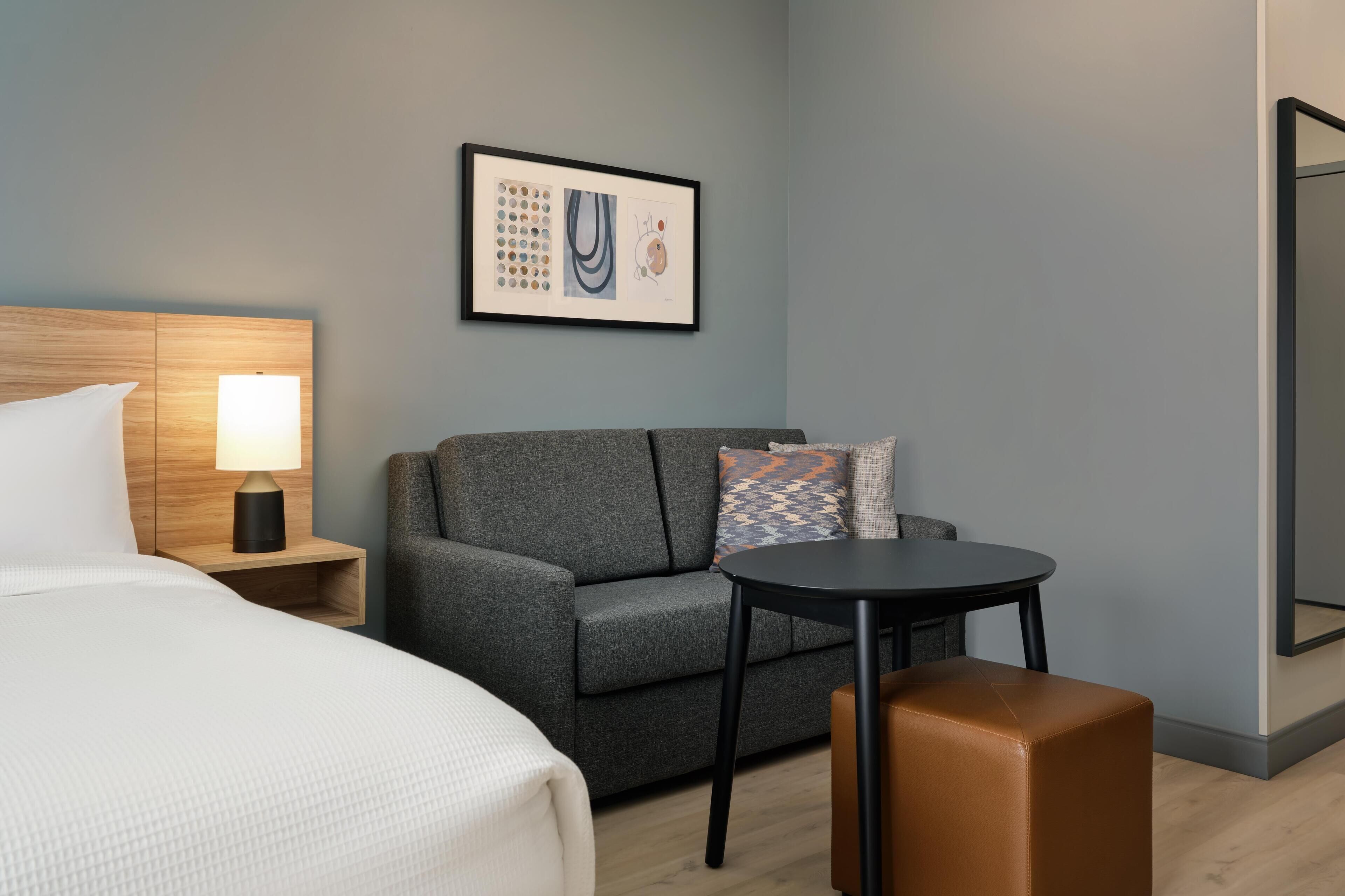 A hotel room with a bed, a gray sofa, a small round table, a nightstand with a lamp, and a framed artwork on the wall.