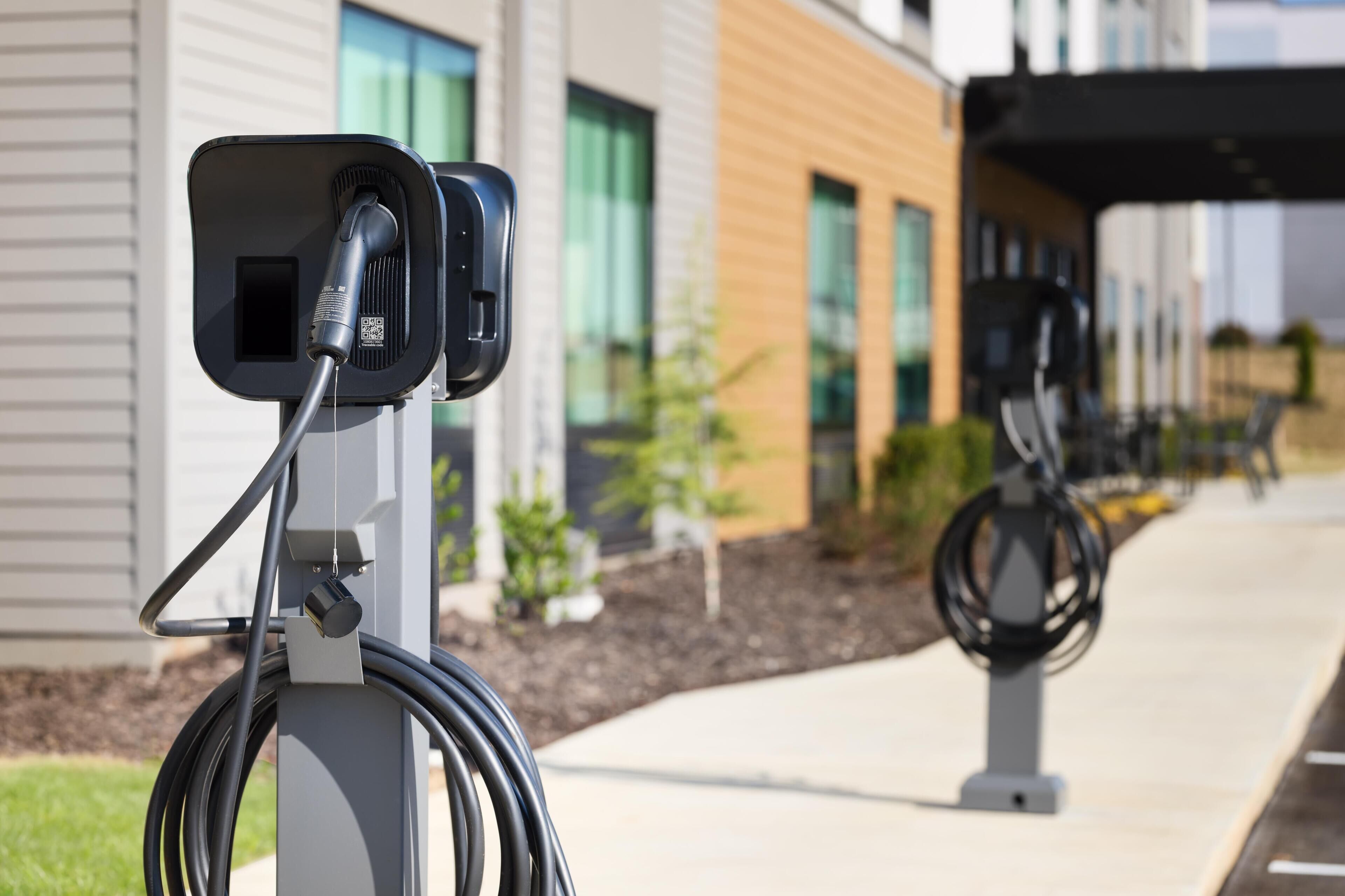 Estação de carregamento para veículos elétricos com cabo conectado, em frente a fachada moderna de um edifício com janelas amplas e área pavimentada.