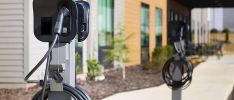 Una estación de carga para vehículos eléctricos con un cable conectado, frente a una fachada de edificio moderno con ventanas amplias y un área pavimentada.