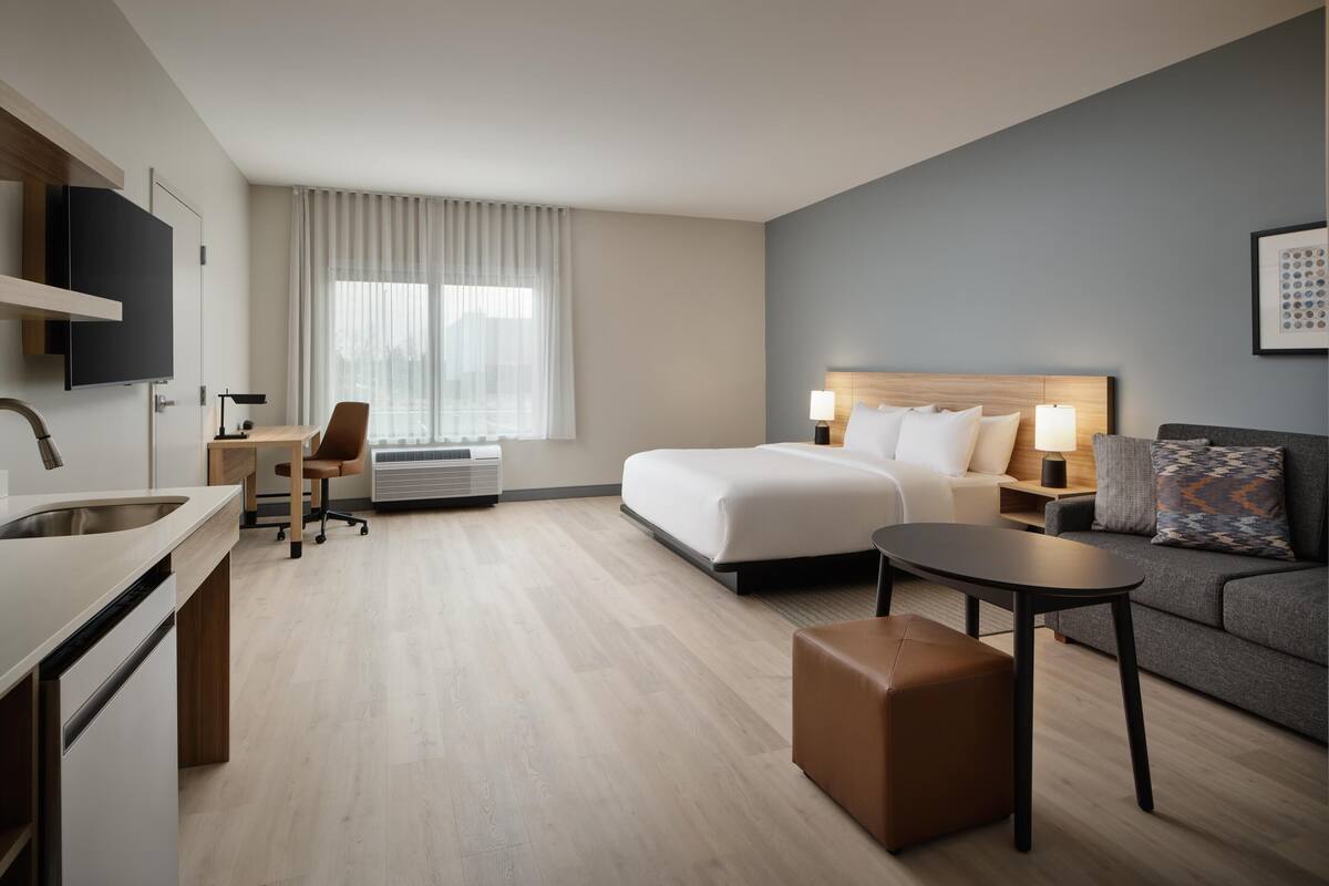 Une chambre d’hôtel moderne équipée d’un lit, d’un canapé, d’un bureau avec une chaise, d’une petite table et d’un téléviseur fixé au mur.