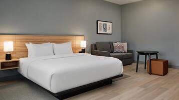 A modern hotel room with a large bed, two bedside tables with lamps, a grey sofa, a small round table, and a brown ottoman.