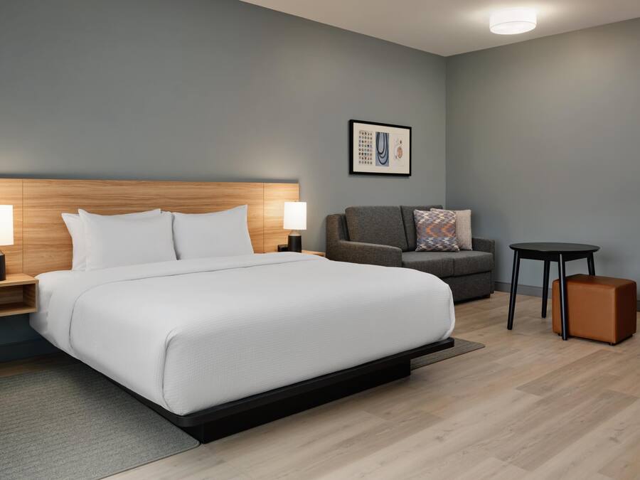 A modern hotel room with a large bed, two bedside tables with lamps, a grey sofa, a small round table, and a brown ottoman.