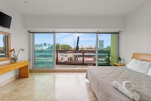 A modern hotel room with a large bed, a wooden desk, a balcony with a view of palm trees and buildings, and a television mounted on the wall.