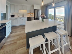 A modern kitchen with white cabinets, a central island with stools, a refrigerator, and a view of the outdoors through large windows. - Kirk's Mema's Beach by Topsail Realty (Holly Ridge)