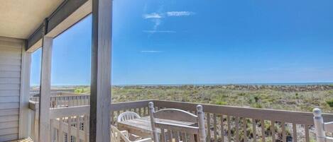 A wooden deck with a table and chairs, overlooking a beach with sparse vegetation.