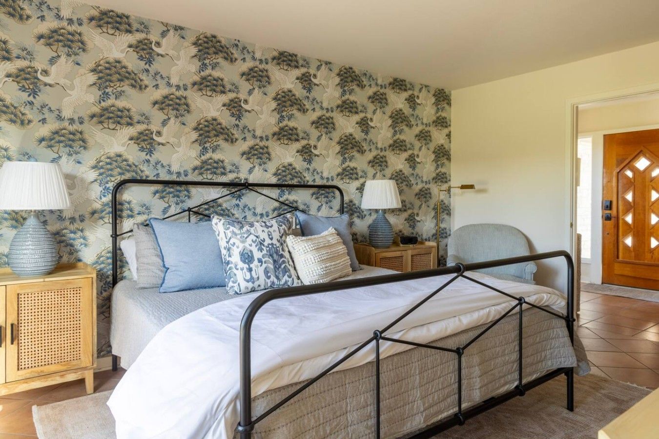 A bedroom with a metal bed frame, two bedside tables with lamps, a rattan cabinet, and a patterned wallpaper.
