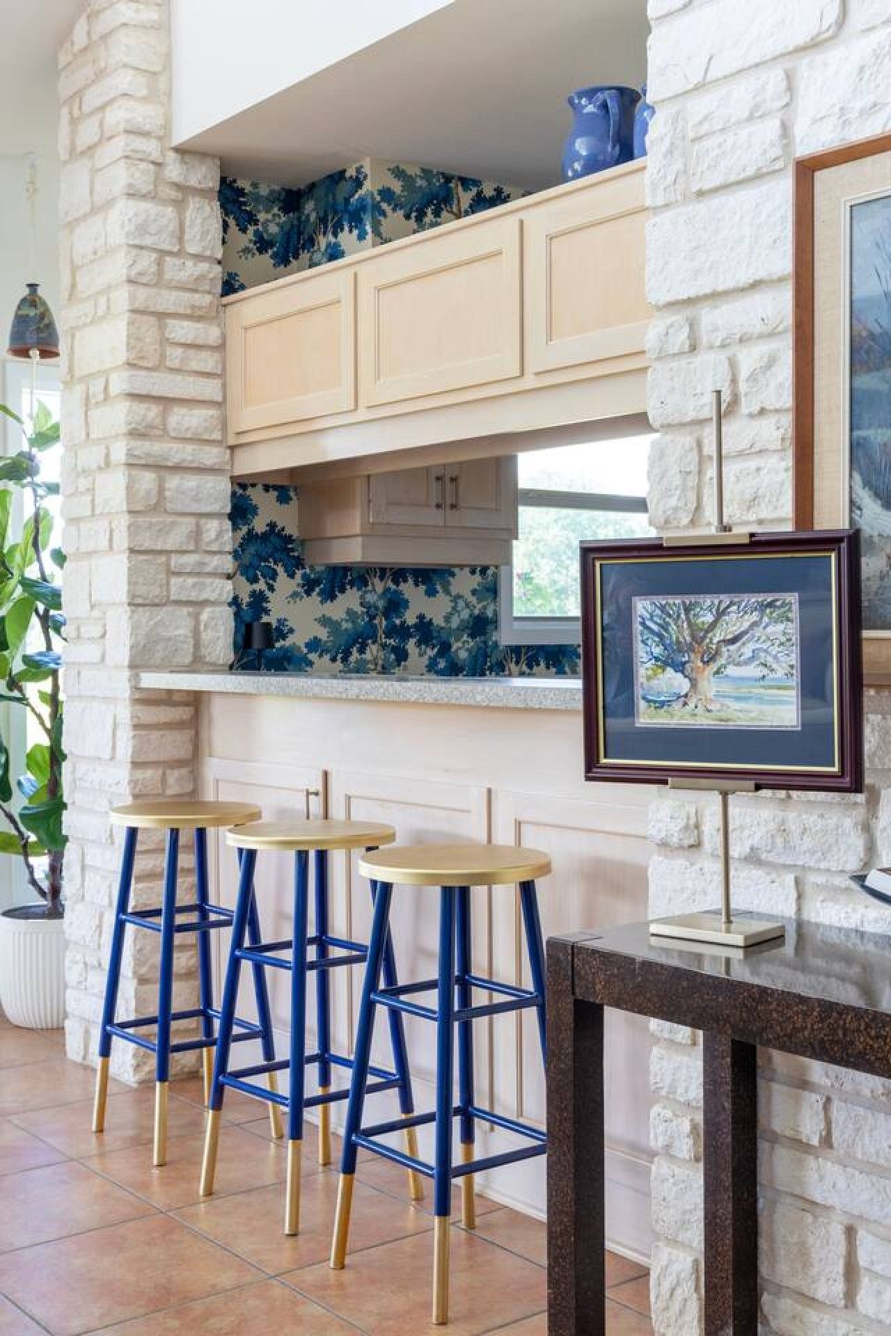A kitchen with white stone walls, blue floral backsplash, and wooden cabinets. There are three blue bar stools and a framed picture on the wall.