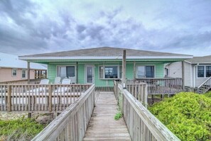 Uma casa térrea com fachada verde, um deck de madeira e uma varanda coberta com cadeiras.