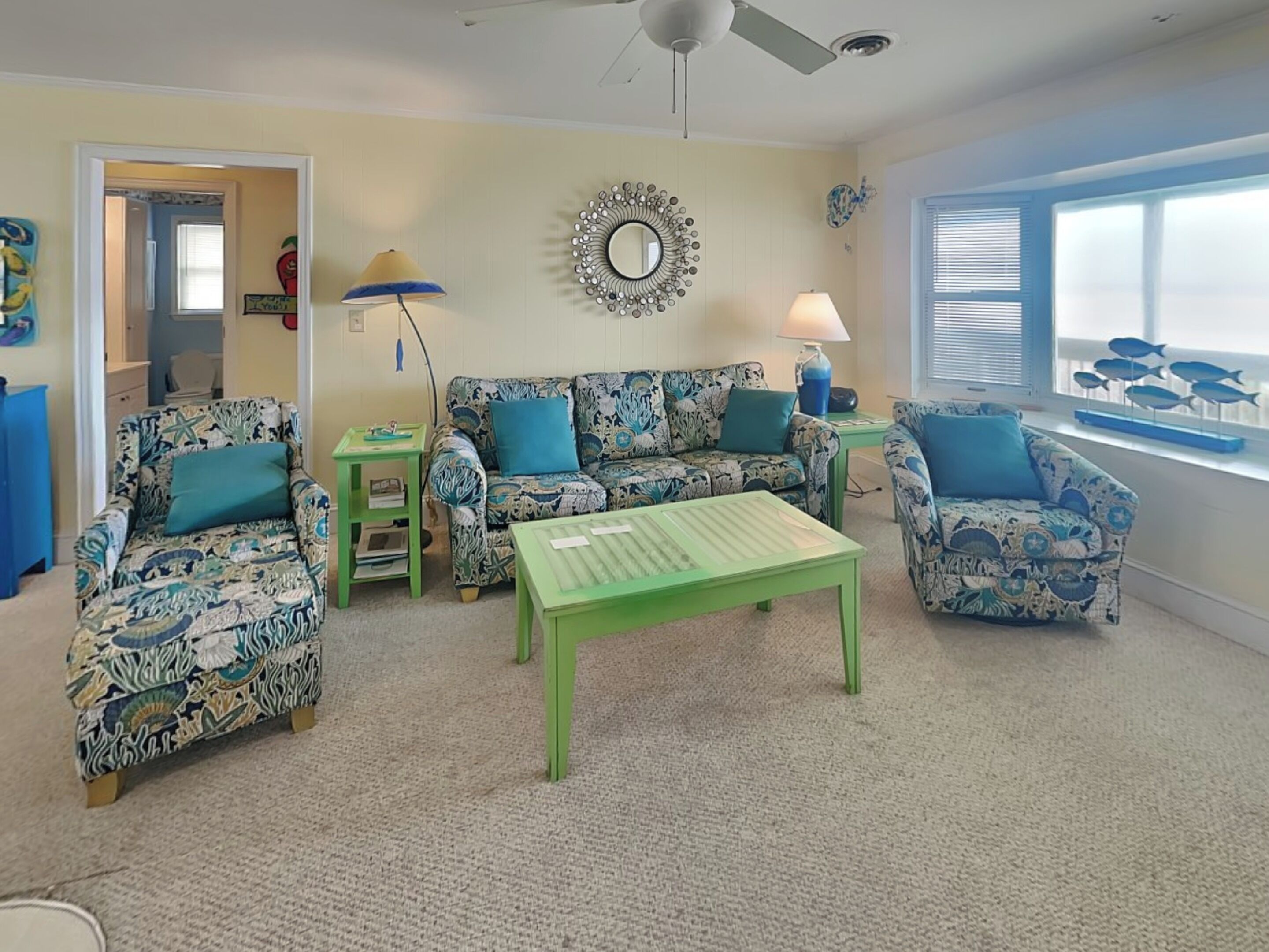 A living room with patterned furniture, a ceiling fan, a decorative mirror, and a view of the sea through a window.