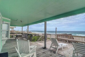 Área externa coberta com deck de madeira, cadeiras Adirondack brancas e vista para o mar.