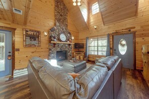 Interior de una cabaña de troncos con chimenea de piedra, sofá de cuero y televisor.