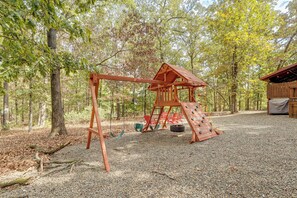 A wooden swing set with a climbing wall and tire swings in a wooded area. - Near Choctaw Landing | 2 King Bedrooms | Game Loft (Broken Bow)