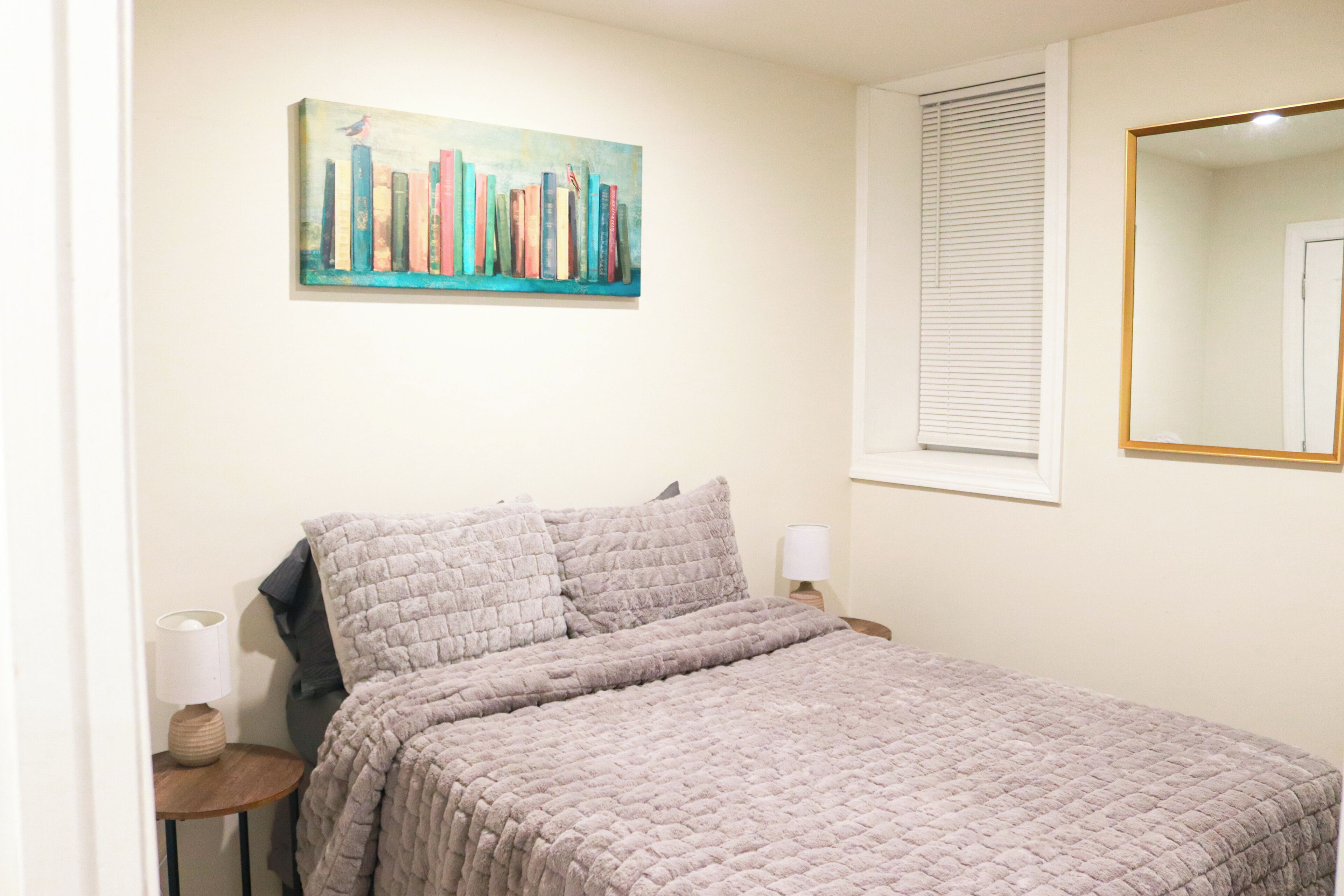 Un dormitorio con una cama, un cuadro de libros, una ventana con persianas y un espejo.