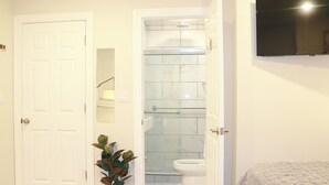 Un baño con puerta blanca, ducha con puertas de cristal, inodoro y televisor montado.