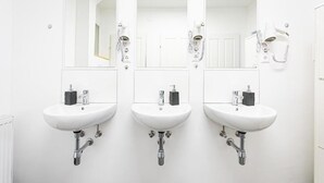 Un baño con tres lavamanos blancos, cada uno con un espejo encima. Hay secadores de pelo montados en la pared a la derecha.