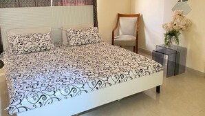 Un dormitorio con una cama, una silla, una mesita con un florero y un adorno de pared.
