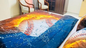 Habitación con una cama cubierta con una colcha grande y colorida de temática de béisbol, dos sillas y una puerta de madera.