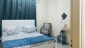 Un dormitorio con una colcha estampada, una silla, una mesita con una planta y un adorno en la pared.