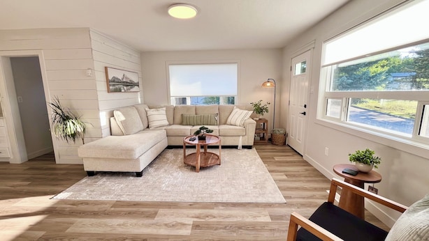 A modern living room with a sectional sofa, a round coffee table, and a view of the outdoors through large windows. - Alpine Escape-Completely Renovated Home, 20 minutes from Jasper Park Gates (Hinton)